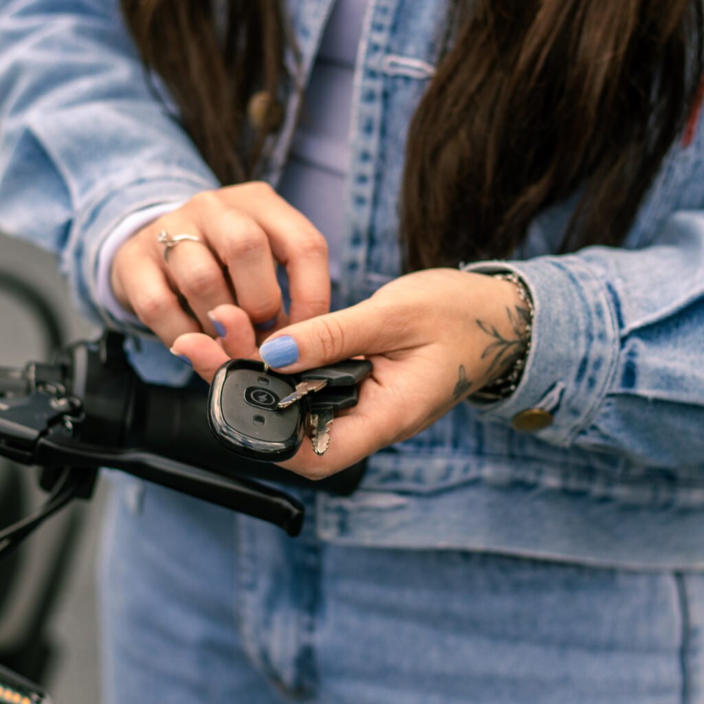 Bicicleta Elétrica no Inverno: Guia Completo Para Pedalar no Frio com Segurança 3 Foto em close-up das mãos de uma mulher segurando um chaveiro da Two Dogs. Suas unhas estão pintadas de azul claro. Uma das mãos, que tem uma tatuagem de raminho no pulso e um anel, segura um controle remoto preto (chave presencial) e duas chaves de metal presas a ele. A outra mão está prestes a manusear as chaves. Ela veste uma jaqueta jeans azul sobre uma camisa de cor clara.
