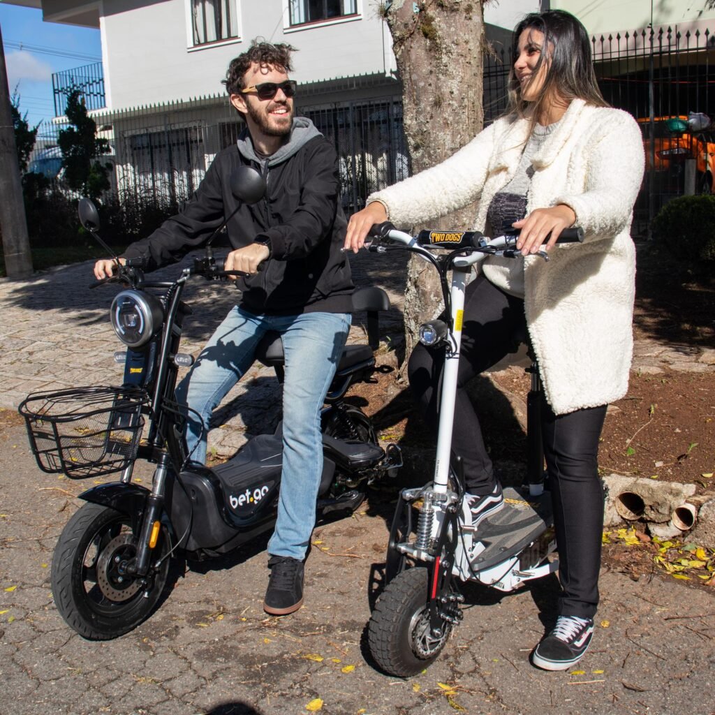 Bicicleta Elétrica no Inverno: Guia Completo Para Pedalar no Frio com Segurança 2 Foto de um casal sorrindo sobre veículos elétricos da Two Dogs em uma rua residencial num dia ensolarado. O homem, à esquerda, usa óculos de sol, jaqueta preta e calça jeans, sentado em uma scooter elétrica preta da Two Dogs. A mulher, à direita, sorri usando um casaco fofo branco e calça preta, em pé sobre um patinete elétrico branco da Two Dogs. Eles estão próximos a uma árvore e edifícios ao fundo.