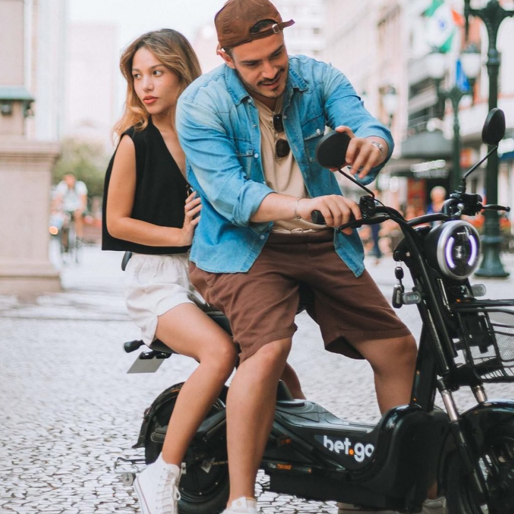 Casal em uma bicicleta elétrica Bet.Go preta da Two Dogs, em uma rua de pedras no centro da cidade, com prédios históricos ao fundo.