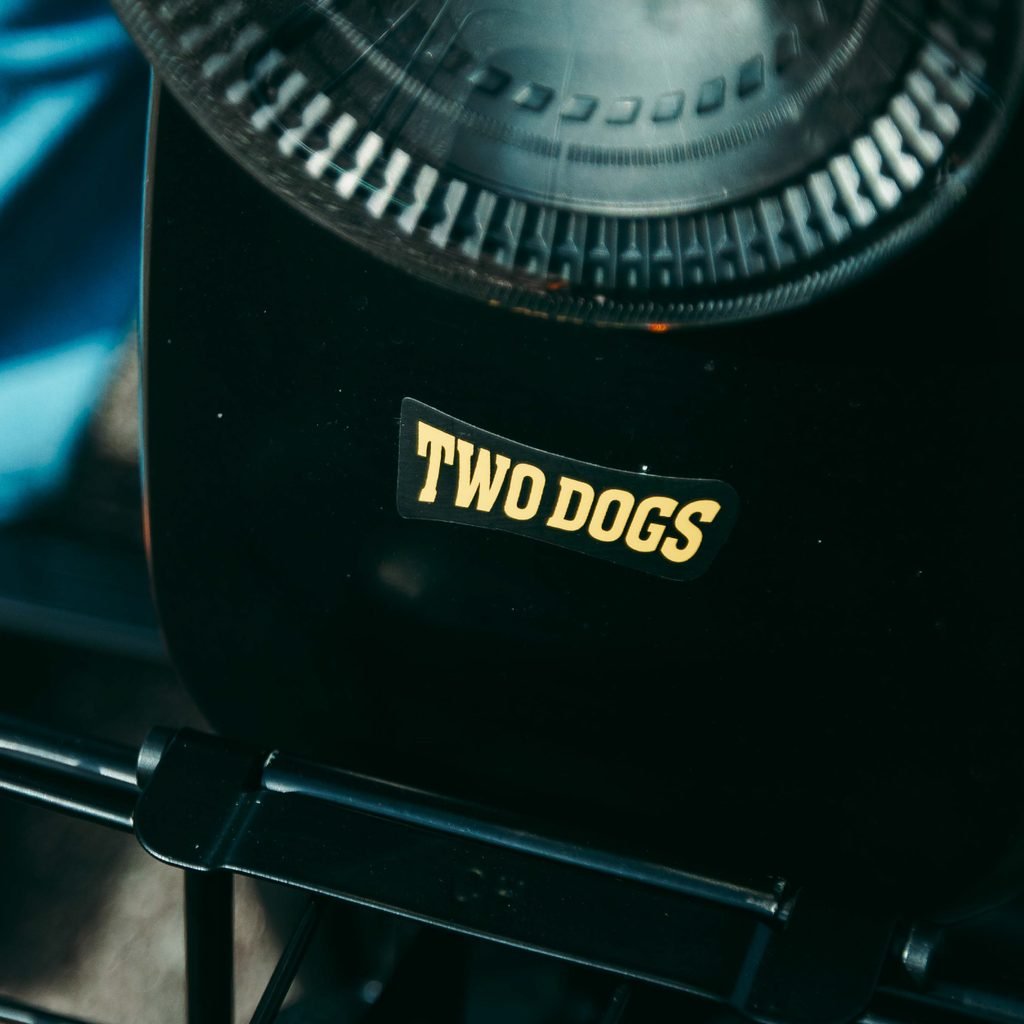 Close-up do logotipo amarelo da Two Dogs na parte frontal de uma bicicleta elétrica preta, com destaque para o acabamento do equipamento.